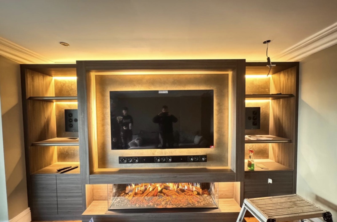 Bespoke media wall in smoked oak with a gold-leaf television surround, three-sided glass fire below and LED-lit display shelving either side