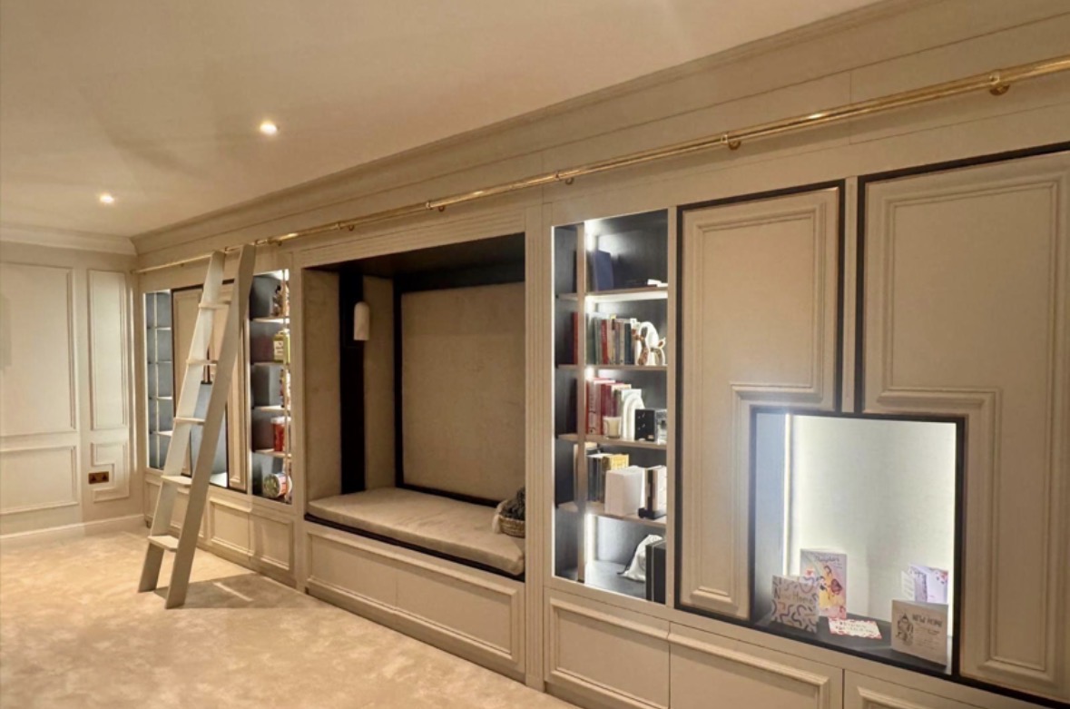 Hand-painted panelled library wall with an upholstered reading nook, LED-lit bookshelves, cupboards below and a brass rail with a sliding ladder