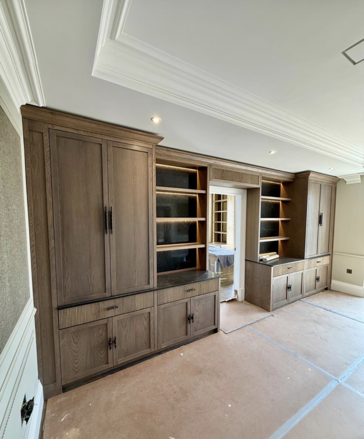 Full-wall run of smoked oak living room cabinetry with panelled doors, fluted drawer fronts, dark stone tops and LED-lit shelving either side of a doorway