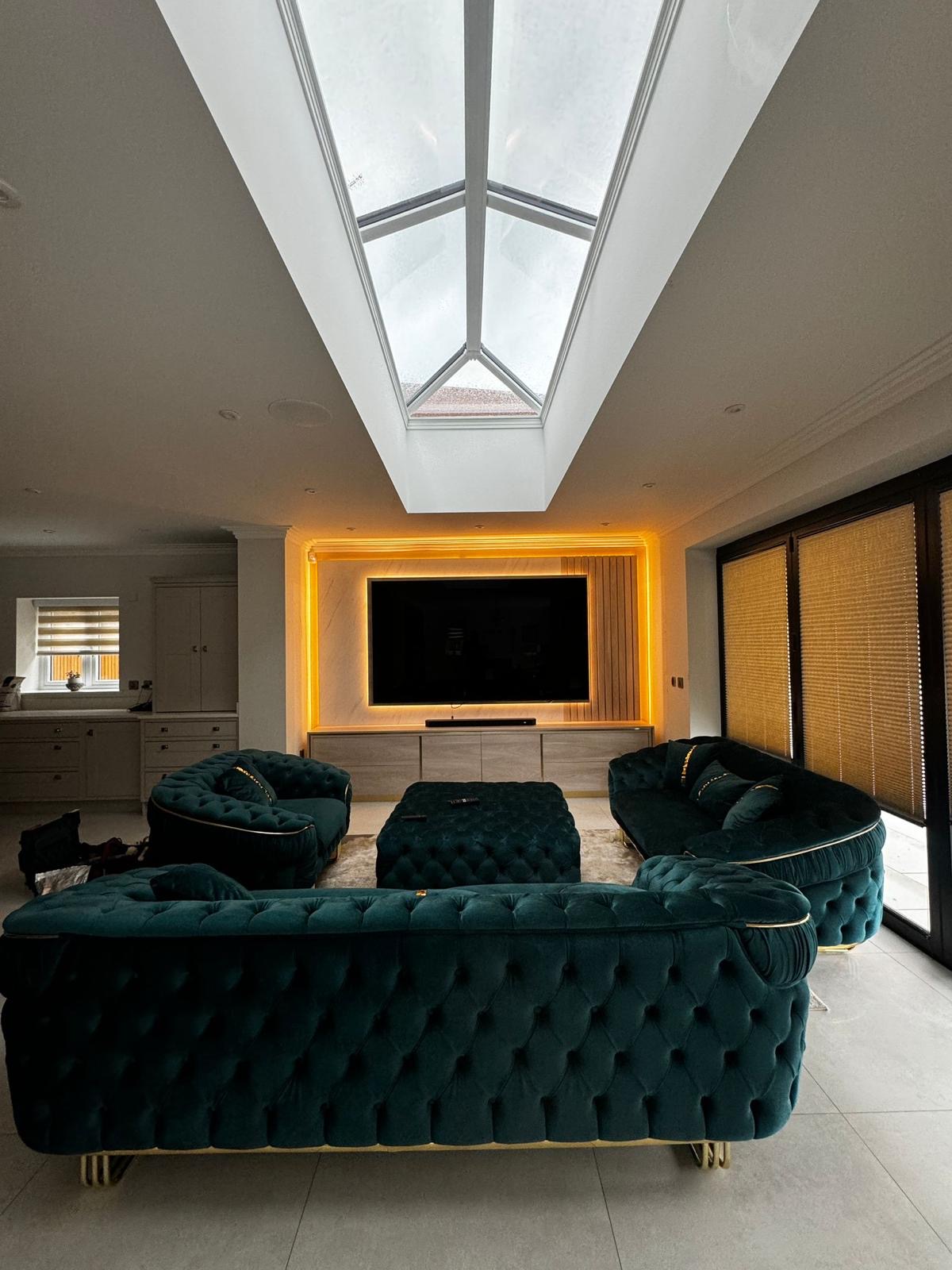 Full-height fluted media wall with an inset fire, LED-lit display shelving and green velvet sofas beneath a skylight